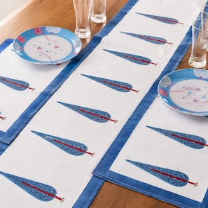 May include: A white table runner and matching placemats featuring a repeating blue leaf pattern with red stems and blue borders. The set is arranged with two round plates decorated with pink flowers and blue accents, plus two clear drinking glasses.