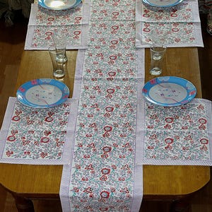 Peut inclure: Une table en bois avec un chemin de table, des sets de table et des assiettes à motifs floraux. Le tissu blanc présente des fleurs rouges et des feuilles vertes. Des assiettes à bord bleu et des verres transparents complètent l'ensemble.