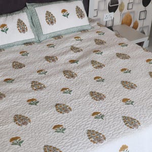May include: A white quilted bedspread and two pillowcases with a floral pattern in shades of brown, yellow, and green. The pillowcases have a green striped border. The bedspread has a repeating pattern of stylized flowers and paisley shapes.