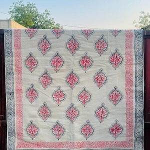 Puede incluir: Un textil color crema con un patrón repetido de diseños florales rojos y azul marino. El borde presenta un patrón rojo y azul marino con flecos en el borde inferior. El textil está colgado al aire libre.