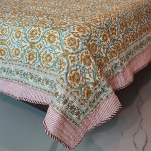Peut inclure: Une couverture de lit à motifs avec un design floral dans les tons or, turquoise et blanc. La bordure présente un motif à rayures roses et blanches. La couverture est pliée, révélant l'imprimé et la texture détaillés.