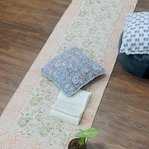 Può includere: Un tappeto rettangolare color crema con un motivo floreale verde e pesca. Il tappeto ha un bordo color pesca con un design dettagliato e frange alle estremità. Due cuscini decorativi e un libro aperto poggiano sul tappeto.