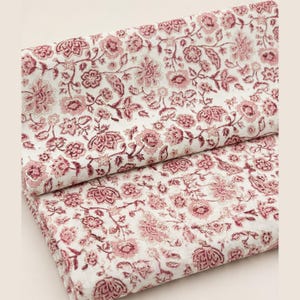 Peut inclure: Un morceau de tissu plié présentant un motif floral répétitif rouge foncé sur fond blanc. Le textile affiche des illustrations botaniques complexes de diverses fleurs, feuilles et vignes réparties uniformément sur toute la surface.