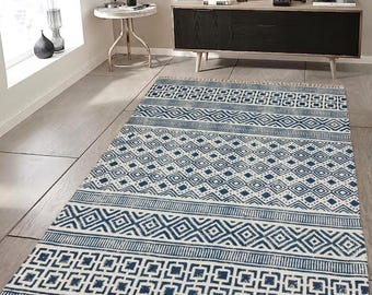 May include: A rectangular area rug featuring a repeating pattern of blue geometric shapes, including diamonds and squares, on a cream-colored background. The rug has a distressed texture and is finished with short, light-colored fringe on both ends.