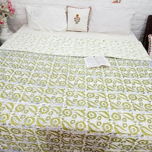 Puede incluir: Una colcha de algodón con un patrón repetitivo de estampado en bloque de flores y cachemir en verde y blanco. La colcha está extendida sobre una cama con dos almohadas blancas y un cojín central con motivo floral. Un libro abierto descansa encima.