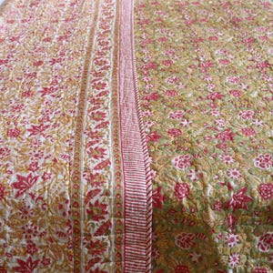 Op de afbeelding: Een quilt met bloemenpatroon met een crèmekleurige achtergrond en rode en gouden bloemen. De quilt heeft een rood-witte gestreepte rand en een bloemenrand met rode bloemen en gouden accenten.