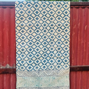 Puede incluir: Una alfombra rectangular con un diseño de diamantes repetido en azul y crema. El borde inferior presenta un patrón geométrico en verde y azul, con flecos. La alfombra cuelga contra una superficie de metal corrugado rojo.