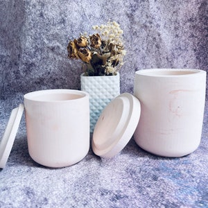 May include: Two white ceramic candle jars with lids. The jars have a smooth, matte finish. One jar is open and the lid is placed next to it. The other jar is closed. The jars are sitting on a gray textured surface.