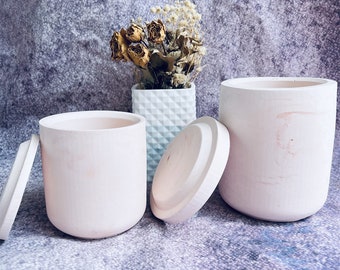 May include: Two white ceramic candle jars with lids. The jars have a smooth, matte finish. One jar is open and the lid is placed next to it. The other jar is closed. The jars are sitting on a gray textured surface.
