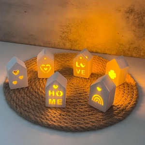 Puede incluir: Seis casas blancas de cerámica con diferentes palabras y diseños recortados. Las casas están iluminadas desde el interior con luces LED blancas cálidas. Las casas están dispuestas sobre una alfombra de yute tejida.