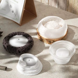 Könnte beinhalten: Vier verschiedene Kerzenhalter, jeder mit einem einzigartigen Design. Der erste ist ein schwarzer, strukturierter Halter mit einer weißen Kerze. Der zweite ist ein weißer, runder Halter mit einer weißen Kerze. Der dritte ist ein weißer, runder Halter mit einer weißen Kerze und einem Holzsockel. Der vierte ist ein weißer, strukturierter Halter mit einer weißen Kerze.