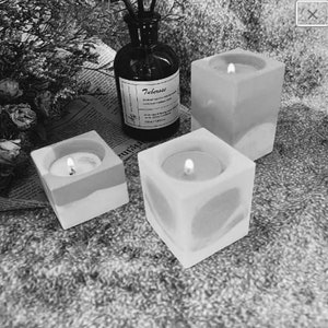 May include: Three white square candle holders with a textured surface. Each holder has a small, round, white candle inside. The candles are lit.