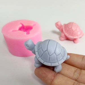 May include: A light grey turtle figurine with a textured shell, next to a pink turtle and a pink silicone mould. The mould has a turtle shape. The turtle figurine is held in a hand.