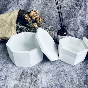 May include: Two white octagonal boxes with lids. The boxes are made of a smooth, white material and have a slightly textured surface. The lids are slightly ajar, revealing the empty interiors of the boxes.