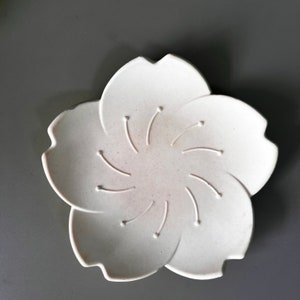 Può includere: Piatto in ceramica bianca a forma di fiore con cinque petali. Il piatto ha un design in rilievo al centro che ricorda un fiore.