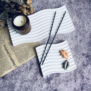 May include: Two white ceramic trays with wavy edges. One tray has two black incense sticks and the other has two small decorative objects shaped like lips.