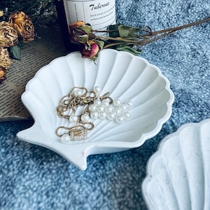 May include: Two white ceramic seashell-shaped dishes, one with a gold chain necklace and pearl beads, the other empty. The dishes are on a blue patterned surface.