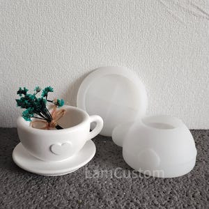 May include: White silicone mould for making a teacup and saucer with a heart design. The mould is shown with a small bouquet of green flowers in the teacup.