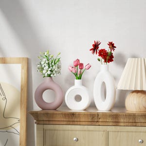 May include: Three ceramic vases in different shapes and colors. The vase on the left is pink and has a round opening. The middle vase is white with a circular opening. The vase on the right is white with a vertical opening. All three vases are sitting on a wooden surface.