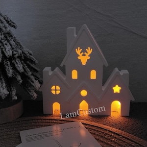 Könnte beinhalten: Weißes Holz-Weihnachtsdorfhaus mit ausgeschnittenen Fenstern und einem Rentier-Design. Das Haus ist mit warmweißen Lichtern beleuchtet. Der Text "LamCustom" ist auf der Basis sichtbar.