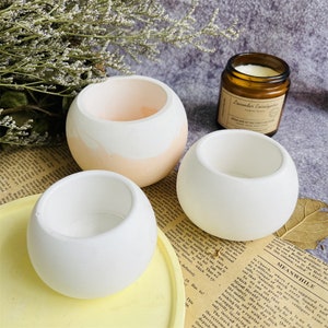 Puede incluir: Tres macetas redondas de hormigón, dos blancas y una con un esquema de color melocotón y blanco. Las macetas están dispuestas sobre una superficie de color amarillo claro.