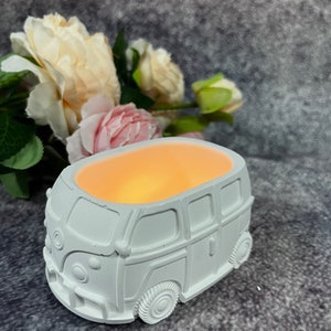 Może przedstawiać: Biała ceramiczna doniczka w kształcie zabytkowego Volkswagena T1 z jasnym pomarańczowym światłem pochodzącym z wnętrza.