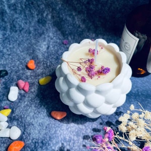 May include: White ceramic candle holder with a textured, bubble-like design. The holder contains a white candle with a single wick and dried purple flowers.