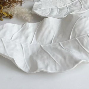 May include: Two white ceramic leaf-shaped serving dishes. The dishes are made to look like large leaves with a textured surface.