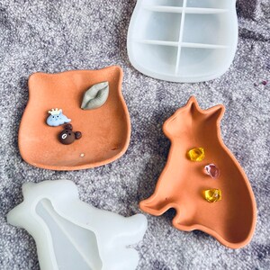 May include: Three terracotta-colored resin trays in the shapes of an owl, a cat, and a bear. The owl tray has a small cloud with a crown, a bear, and a lip charm. The cat tray has three clear glass gems. The bear tray is empty.
