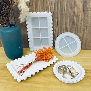 May include: A selection of white silicone moulds in different shapes, including a rectangular tray, a circular tray, and window-shaped designs. The trays have a scalloped edge. A small bouquet of dried orange flowers and rings are also visible.