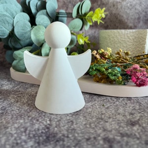 Puede incluir: Figurita de ángel de cerámica blanca con cuerpo en forma de cono y alas. El ángel está de pie sobre una superficie blanca.