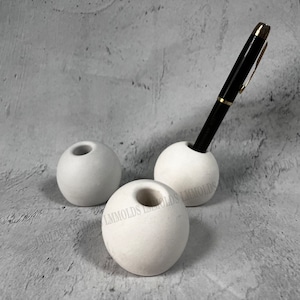 May include: Three white spherical pen holders. One holder contains a black pen with gold accents. The pen holders are made of a smooth, matte material. The pen holders are arranged on a grey surface.