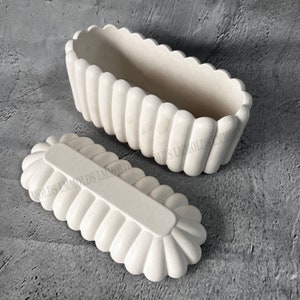 May include: White oval-shaped silicone mold with a ribbed texture. The mold has a matching lid. The mold is designed to create a decorative, ribbed, oval-shaped object.