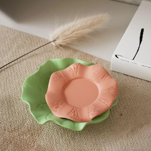 May include: A set of two ceramic coasters in the shape of flowers. The larger coaster is green and the smaller coaster is peach. Both coasters have a scalloped edge.