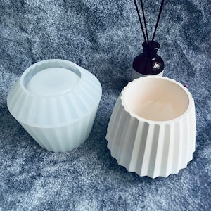 May include: A white, ribbed, geometric planter next to a clear silicone mold of the same shape. The planter has a wide opening and a textured exterior. A dark brown bottle with reeds is in the background.