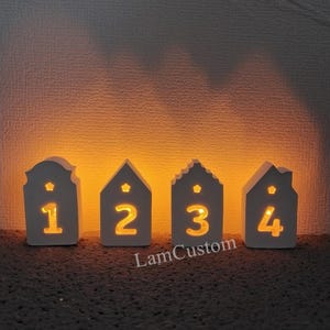 Könnte beinhalten: Vier weiße Holzhäuser mit einem Stern und einer Nummer auf jedem. Die Häuser sind von innen beleuchtet und strahlen ein warmes Licht aus. Die Nummern auf den Häusern sind 1, 2, 3 und 4.