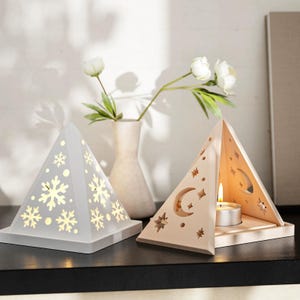 Könnte beinhalten: Zwei pyramidenförmige Kerzenhalter aus Holz. Einer ist weiß mit einem Schneeflockenmuster und der andere ist beige mit einem Mond- und Sternenmuster. Beide haben eine kleine Öffnung für ein Teelicht.
