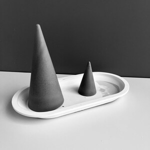 Può includere: Un vassoio ovale in ceramica bianca con due porta anelli a forma di cono in cemento grigio. Il cono più grande è a sinistra e quello più piccolo a destra.