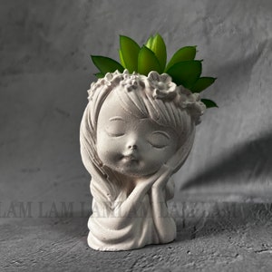 Könnte beinhalten: Weißer Keramik-Pflanztopf in Form eines Mädchenskopfes mit geschlossenen Augen und einer Blumenkrone. Der Topf hat eine Sukkulente, die aus der Oberseite wächst.