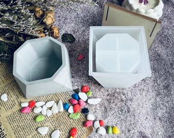 May include: Two silicone molds for making concrete planters. One mold is a geometric shape with a square base and a pointed top. The other mold is a square shape with a flat top. The molds are white and are sitting on a gray surface with colorful pebbles.