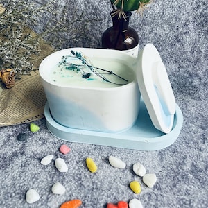 May include: A white and blue oval concrete candle holder with a lid. The candle holder has a light blue and white ombre design. The candle holder is filled with a white candle and dried flowers.