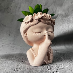 May include: A white ceramic planter shaped like a girl's bust with a flower crown and her finger to her lips. The planter has a green succulent plant growing out of the top.