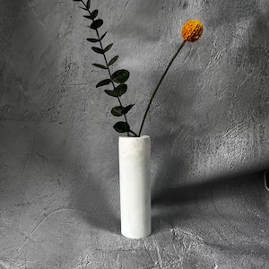 May include: A white cylindrical vase with a single stem of green leaves and a yellow flower.