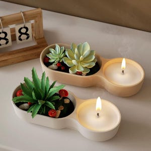May include: Two white and beige ceramic candle holders with a double design. Each holder has a small planter with artificial succulents and a candle. One candle is lit.