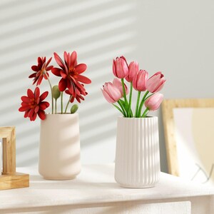 May include: Two ceramic vases with different floral arrangements. The vase on the left is a light pink color and holds a bouquet of red flowers. The vase on the right is white with vertical ridges and holds a bouquet of pink tulips.
