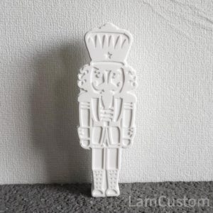 Pode incluir: Cortador de biscoitos de plástico branco em forma de quebra-nozes tradicional. O quebra-nozes tem uma coroa, um bigode e um uniforme detalhado.