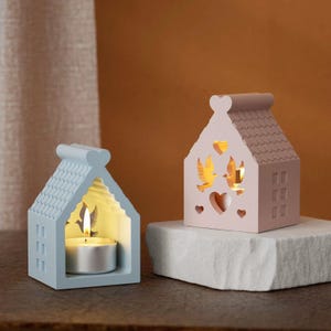 Puede incluir: Dos portavelas decorativos con forma de casas. La casa de la izquierda es azul claro con una vela blanca en su interior. La casa de la derecha es rosa con recortes de pájaros y corazones.