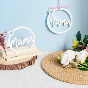 Könnte beinhalten: Zwei weiße Holzschilder mit dem Wort "Mama" in einer kursiv geschriebenen Schrift. Die Schilder hängen an einer Schnur und sind mit einem kleinen Herzen verziert. Die Schilder befinden sich auf einem hellblauen Hintergrund.