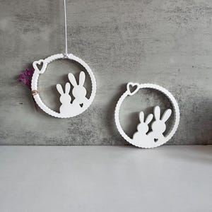 Könnte beinhalten: Zwei weiße Holz Kreise mit Hasensilhouetten und kleinen Herzen. Die Kreise hängen an einer weißen Schnur.