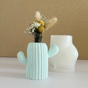 May include: A light blue ceramic cactus-shaped vase with a dried flower arrangement inside. The vase has a ribbed texture and two small arms. The vase is sitting on a white surface.